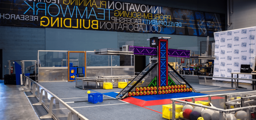 A robotics practice field at Kettering University's FIRST Robotics Community Center, featuring game elements, a central structure, and banners promoting innovation and teamwork.